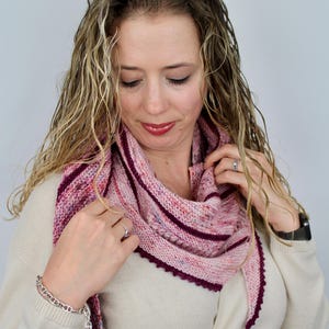 May include: A woman wearing a cream colored sweater with a pink and burgundy striped shawl. The shawl has a scalloped edge and is made of a soft yarn.