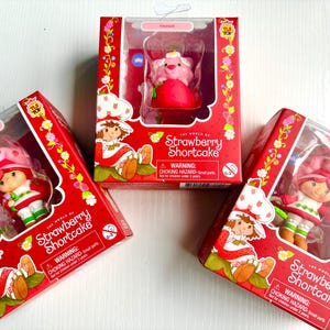 May include: Three boxes of Strawberry Shortcake toys. Each box features a different Strawberry Shortcake character with a red and white background. The boxes have a warning label for choking hazards.