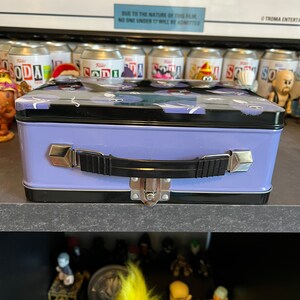 May include: A purple metal lunchbox with a black handle and a latch. The lid has a black and white pattern of cartoon ghosts. The lunchbox is sitting on a shelf with other items.