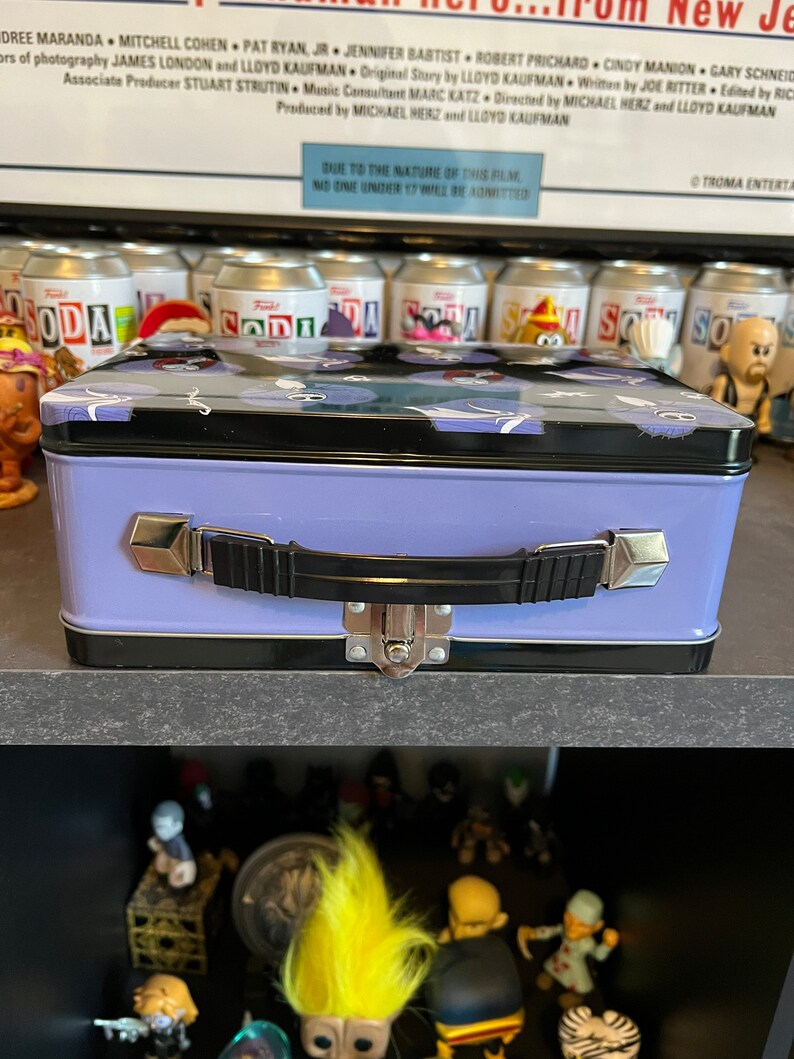 May include: A purple metal lunchbox with a black handle and a latch. The lid has a black and white pattern of cartoon ghosts. The lunchbox is sitting on a shelf with other items.