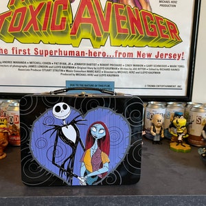 May include: A black metal lunchbox with a heart-shaped design featuring Jack Skellington and Sally from The Nightmare Before Christmas. The lunchbox is sitting on a table with several soda cans and figurines.