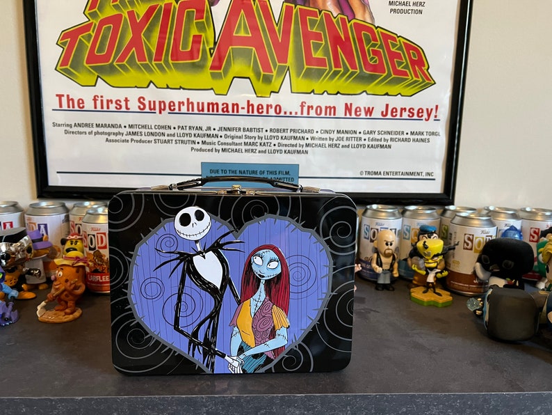 May include: A black metal lunchbox with a heart-shaped design featuring Jack Skellington and Sally from The Nightmare Before Christmas. The lunchbox is sitting on a table with several soda cans and figurines.