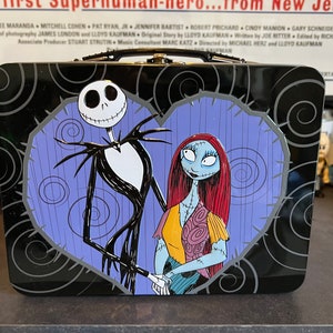 May include: A black metal lunchbox with a purple heart design featuring Jack Skellington and Sally from The Nightmare Before Christmas.