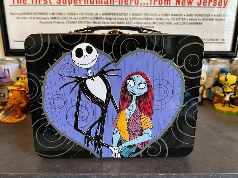 May include: A black metal lunchbox with a purple heart design featuring Jack Skellington and Sally from The Nightmare Before Christmas.