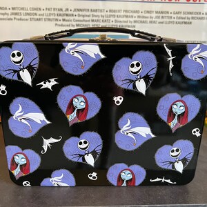 May include: A black metal lunchbox with a handle. The lunchbox has a repeating pattern of purple hearts with images of Jack Skellington and Sally from The Nightmare Before Christmas.