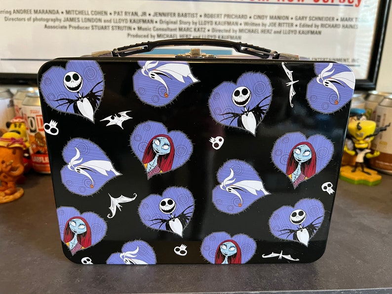 May include: A black metal lunchbox with a handle. The lunchbox has a repeating pattern of purple hearts with images of Jack Skellington and Sally from The Nightmare Before Christmas.