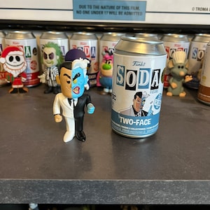 May include: A Funko Soda figure of Two-Face from Batman. The figure is in a can with a black and white design. The can has the words "Funko Soda" and "Two-Face" on it.