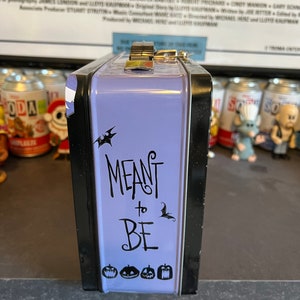 May include: Purple metal lunchbox with a black trim. The lunchbox has a black and white graphic of bats and the text "MEANT to BE". There are also four black and white pumpkin graphics on the bottom of the lunchbox.