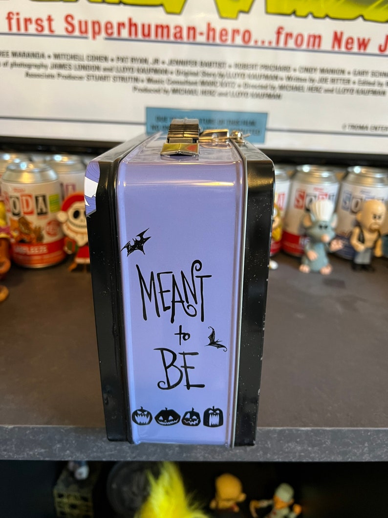 May include: Purple metal lunchbox with a black trim. The lunchbox has a black and white graphic of bats and the text "MEANT to BE". There are also four black and white pumpkin graphics on the bottom of the lunchbox.