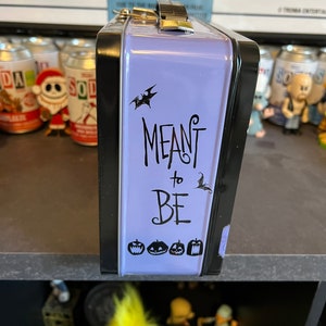 May include: Purple metal lunchbox with a black trim and a black and white graphic that says "MEANT to BE" with a bat and pumpkins.