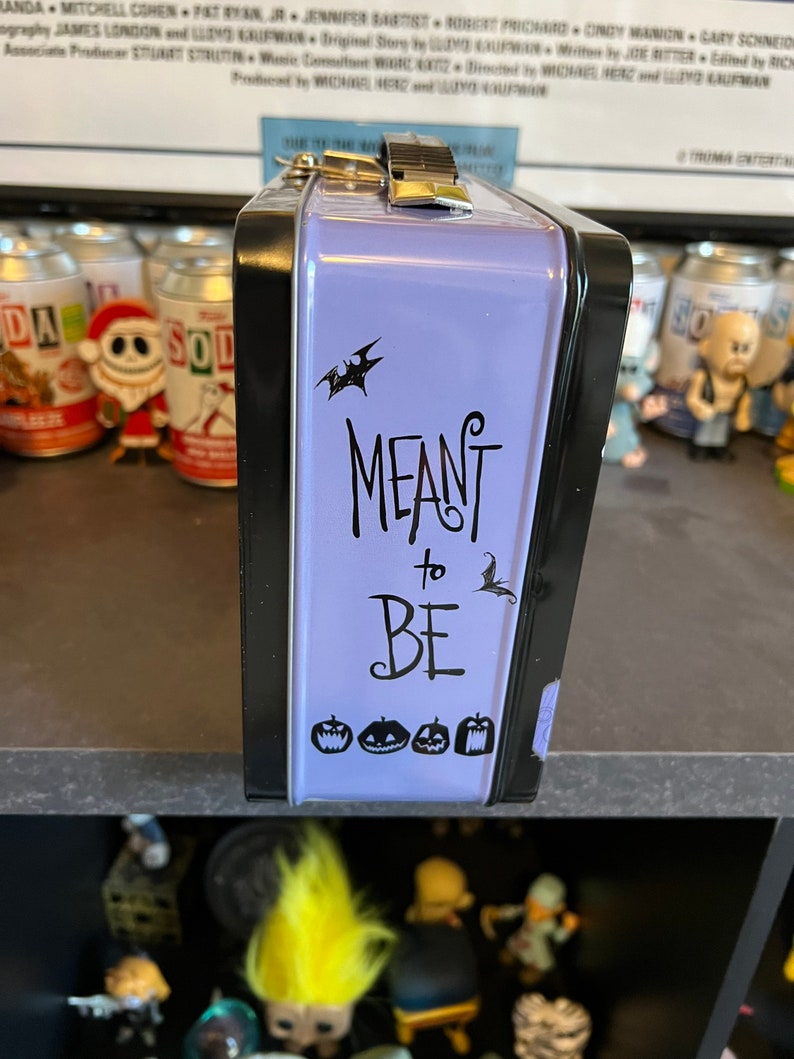 May include: Purple metal lunchbox with a black trim and a black and white graphic that says "MEANT to BE" with a bat and pumpkins.