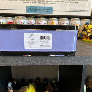 May include: A purple metal lunchbox with a black trim. The front of the box has a white label with the text "Disney's Nightmare Before Christmas" and "Bioworld".