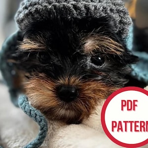 May include: A small puppy with black and brown fur wearing a grey crocheted hat and blue ear coverings. The puppy is nestled in a soft, white blanket. A red circle with the words "PDF PATTERN" is in the lower right corner.