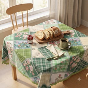 Puede incluir: Un mantel cuadrado con un diseño de patchwork de cuadros verdes, damero crema y motivos florales con rosas rosadas. Los bordes festoneados cubren una mesa de madera con una tabla de cortar con pan rebanado, un tarro de mermelada y una taza.