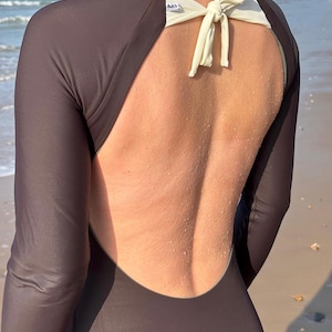 May include: A long-sleeved, dark brown one-piece swimsuit featuring an open back design. The garment includes a cream-colored tie closure at the nape of the neck and a small white brand label visible near the upper back strap area.