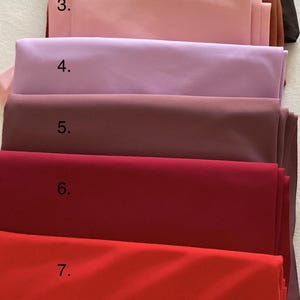 May include: A stack of eight folded fabric swatches labeled 1 through 8, featuring a spectrum of colors including dark brown, rust, light pink, lavender, mauve, deep red, bright red, and orange, all with a smooth, matte, solid-colored finish.