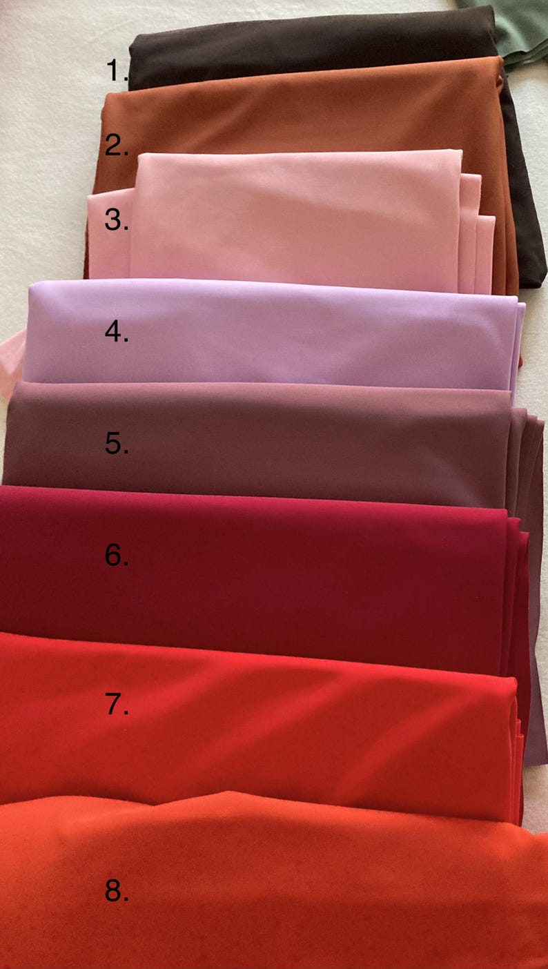 May include: A stack of eight folded fabric swatches labeled 1 through 8, featuring a spectrum of colors including dark brown, rust, light pink, lavender, mauve, deep red, bright red, and orange, all with a smooth, matte, solid-colored finish.