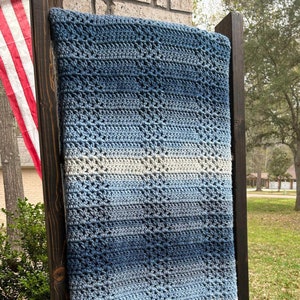 Pode incluir: Uma manta de crochê azul e branca com um padrão de diamante. A manta está pendurada em uma moldura de madeira.