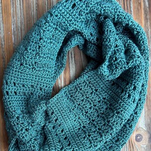 May include: A teal crochet scarf with a lacy pattern. The scarf is folded and displayed on a wooden surface.  Digital Crochet Pattern by Measured and Stitched.