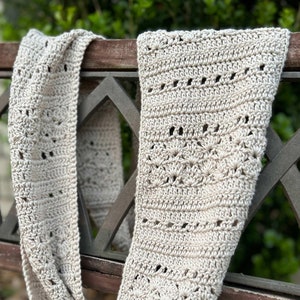 May include: A beige crochet scarf featuring a repeating openwork stitch pattern with horizontal rows of shells and eyelets. The text on the left side reads DIGITAL CROCHET PATTERN by Measured and Stitched in black lettering.