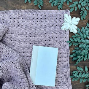May include: A lavender crochet blanket with a textured stitch pattern and small holes. A white book rests on the blanket. The text "DIGITAL CROCHET PATTERN by Measured and Stitched" is visible on the left side of the image.