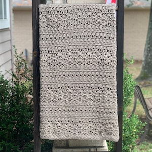 May include: A beige, rectangular, crocheted blanket with a detailed pattern is displayed on a wooden ladder. The blanket's design includes rows of textured stitches and decorative elements. The ladder is positioned outdoors against a brick column.