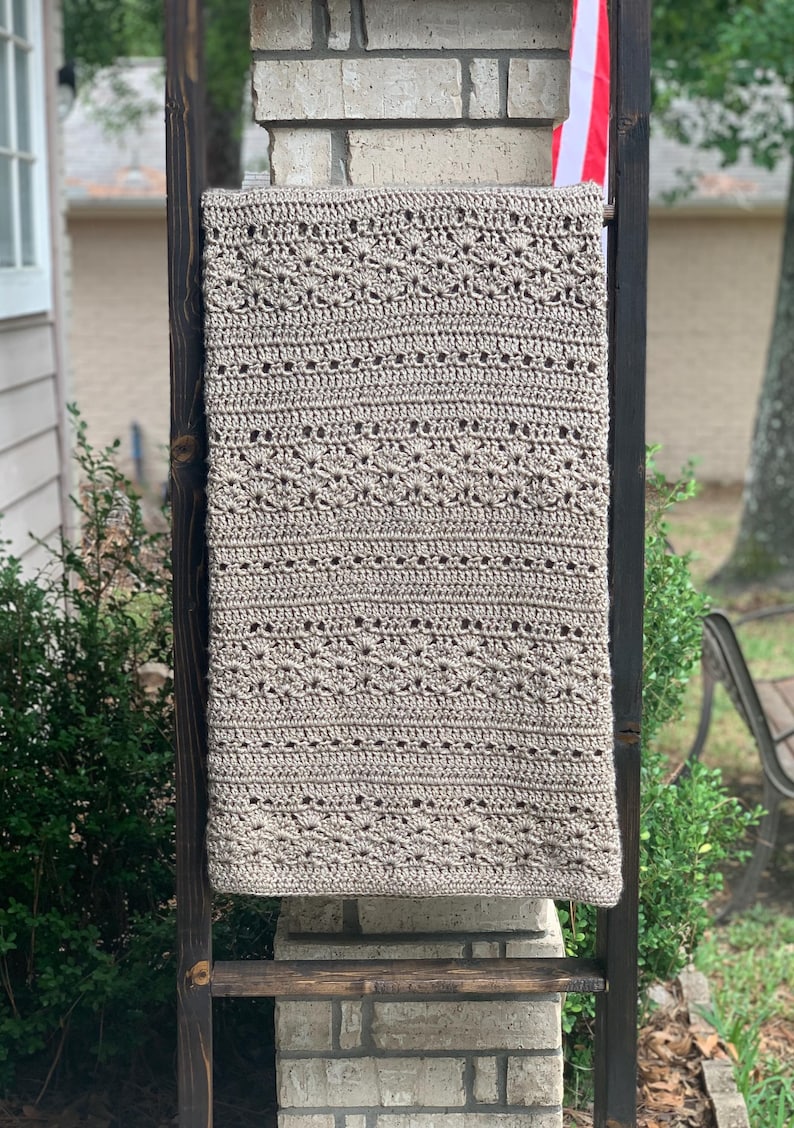 May include: A beige, rectangular, crocheted blanket with a detailed pattern is displayed on a wooden ladder. The blanket's design includes rows of textured stitches and decorative elements. The ladder is positioned outdoors against a brick column.