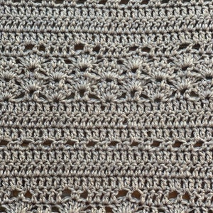 May include: Close-up of a crocheted fabric in a neutral grey colour. The texture features a repeating pattern of openwork and solid stitches, creating a delicate and intricate design. The yarn appears to be a soft, natural fibre.