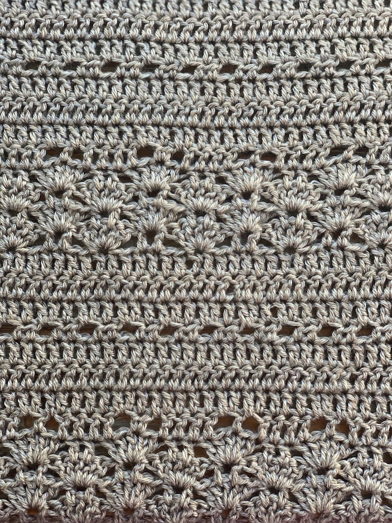 May include: Close-up of a crocheted fabric in a neutral grey colour. The texture features a repeating pattern of openwork and solid stitches, creating a delicate and intricate design. The yarn appears to be a soft, natural fibre.