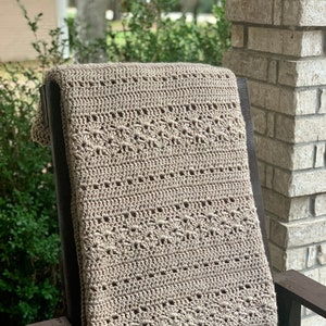May include: A beige crochet blanket with a detailed floral pattern is draped over a dark wooden rocking chair. The blanket's texture and design are clearly visible, showcasing the craftsmanship. The background includes greenery and a brick column.