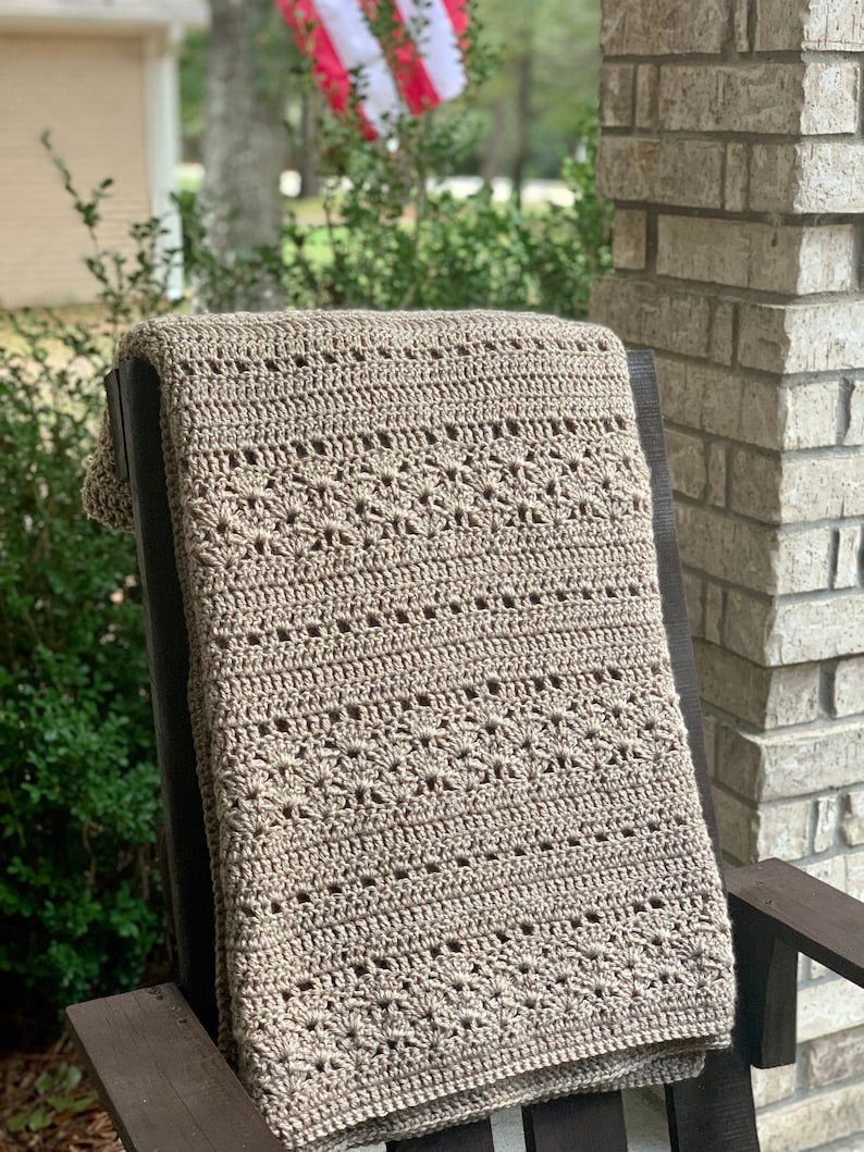 May include: A beige crochet blanket with a detailed floral pattern is draped over a dark wooden rocking chair. The blanket's texture and design are clearly visible, showcasing the craftsmanship. The background includes greenery and a brick column.