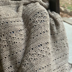 May include: A beige crocheted blanket with a detailed openwork design. The blanket is draped over someone seated in a wooden chair. The texture and colour of the blanket suggest warmth and comfort, suitable for home decor.