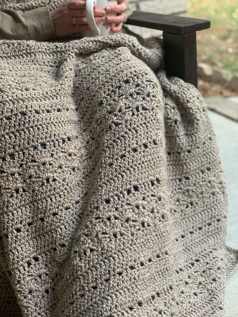 May include: A beige crocheted blanket with a detailed openwork design. The blanket is draped over someone seated in a wooden chair. The texture and colour of the blanket suggest warmth and comfort, suitable for home decor.