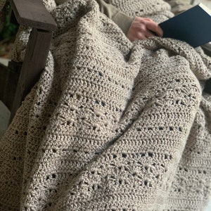 May include: A beige crochet blanket with an openwork design, draped over a wooden chair. The blanket is covering a person holding a book. The neutral colour of the blanket makes it suitable for various interior design schemes.
