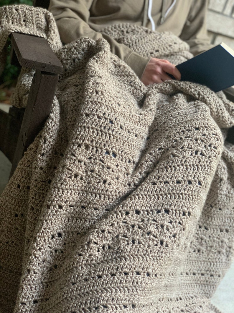 May include: A beige crochet blanket with an openwork design, draped over a wooden chair. The blanket is covering a person holding a book. The neutral colour of the blanket makes it suitable for various interior design schemes.