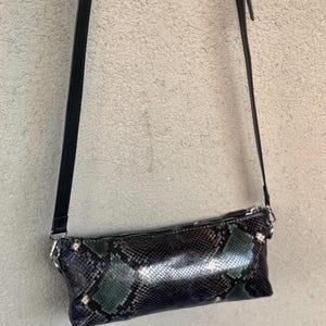 May include: A black and green snakeskin-patterned crossbody bag with a long, black strap. The bag has a rectangular shape with a silver zipper and hardware. The bag is hanging against a neutral background.