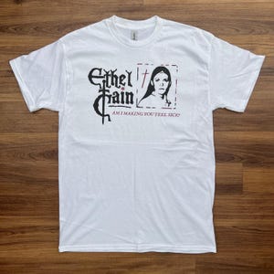 May include: White t-shirt featuring a black graphic with the text "Ethel Cain" and a portrait of a woman. Below the graphic is the text "AM I MAKING YOU FEEL SICK?". The shirt is made of a soft, comfortable material.