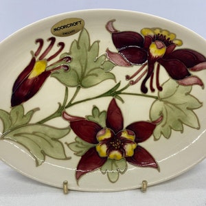 May include: An oval ceramic dish with a white background and a floral design. The flowers are burgundy and yellow with green leaves. The dish has a "Moorcroft England" label.