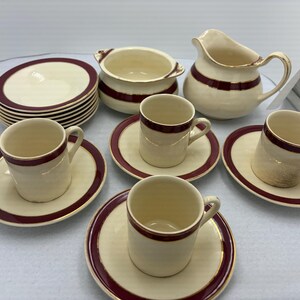 May include: A cream-coloured tea set with burgundy trim. The set includes cups, saucers, a creamer, a sugar bowl, and a stack of plates. The rims of the plates, cups, and saucers are trimmed with a burgundy band.