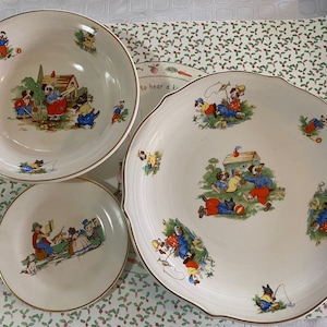 May include: A set of three white ceramic plates with a gold rim. The plates feature a whimsical design of children playing in a garden with a house, trees, and animals. The plates are perfect for serving a child's meal.