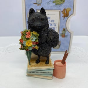 May include: A black fox figurine holding a bouquet of colourful flowers, standing on a stack of books. Beside it is a small pink pot with two brown sticks. The figurine is part of a collection.