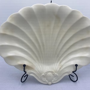 May include: Off-white, shell-shaped decorative plate with a scalloped edge. The plate is supported by a black metal stand. The plate has a detailed, textured surface with a central decorative element.