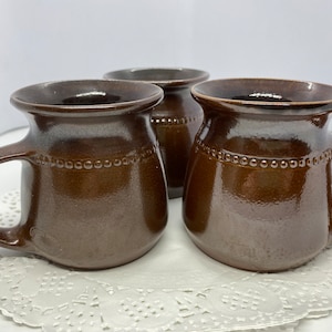 May include: Three brown ceramic mugs with handles. Each mug has a wide, rounded body and a dark brown interior. A band of small, raised dots encircles the upper portion of each mug. The mugs are arranged on a white, lacy doily.