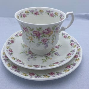May include: A white teacup and saucer set with a floral design. The cup has a handle and sits on a matching saucer, with a smaller plate beneath. The floral design includes pink, yellow, and purple flowers.