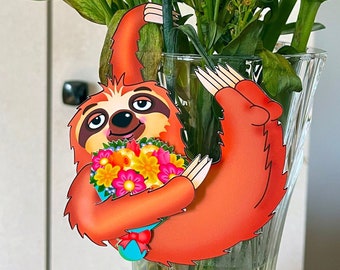 May include: A decorative orange sloth ornament holding a bouquet of colorful flowers, hanging from a clear glass vase filled with orange and red flowers. The sloth has a brown face and is holding a blue bow.