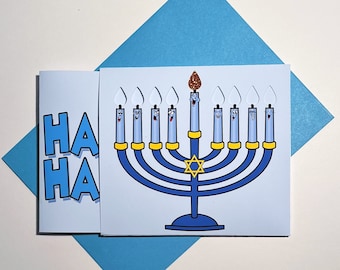 May include: A light blue greeting card with a cartoon menorah design. The menorah has blue candles with smiling faces and a gold Star of David. The card also features the text "HA HA" in blue. The card is on a blue envelope.
