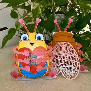 Op de afbeelding: Twee kleurrijke papieren kaarten in de vorm van een kever. Eén is geel met een rood hart met de tekst "Hug Bug". De andere is bruin met een decoratief, opengewerkt ontwerp. Beide hebben roze hartvormige antennes en voeten. De achtergrond is een groene, bladrijke plant.