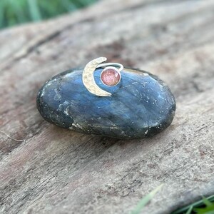 May include: A silver ring with a crescent moon design and a round, pink gemstone. The ring is set on a dark blue and gray stone, with a wooden background and green grass.