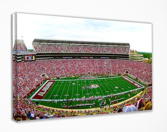 May include: A panoramic view of a packed football stadium with a green field and white yard lines. The stands are filled with fans wearing red and white. The words "Alabama" and "Crimson Tide" are visible on the field.