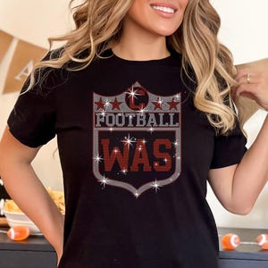 May include: Black t-shirt with a football-themed graphic. The design features the words "FOOTBALL WAS" in a shield shape, with star accents. The shirt is worn by a person in front of a football-themed party setting.